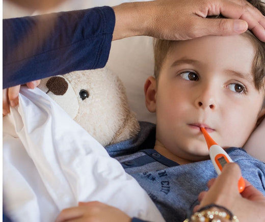 Young boy in bed with a thermometer, cuddling a teddy bear, comforted by his mom's hand on his forehead.
