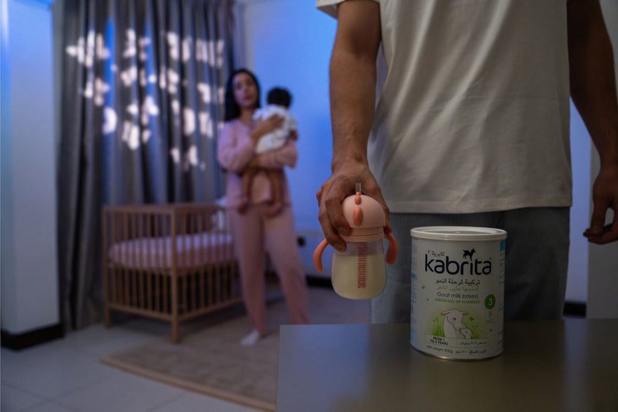 Father preparing Kabrita goat milk formula at night while mother soothes baby in the background – supporting baby sleep routine
